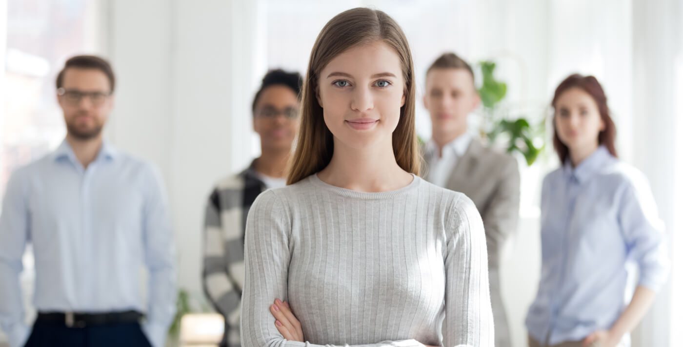 Das Bild zeigt eine Gruppe von Personen in einem Büroraum oder professionellen Umfeld, wobei eine Person im Vordergrund steht und die restlichen Personen im Hintergrund zu sehen sind. Es vermittelt den Eindruck von Teamarbeit oder einem professionellen Treffen.