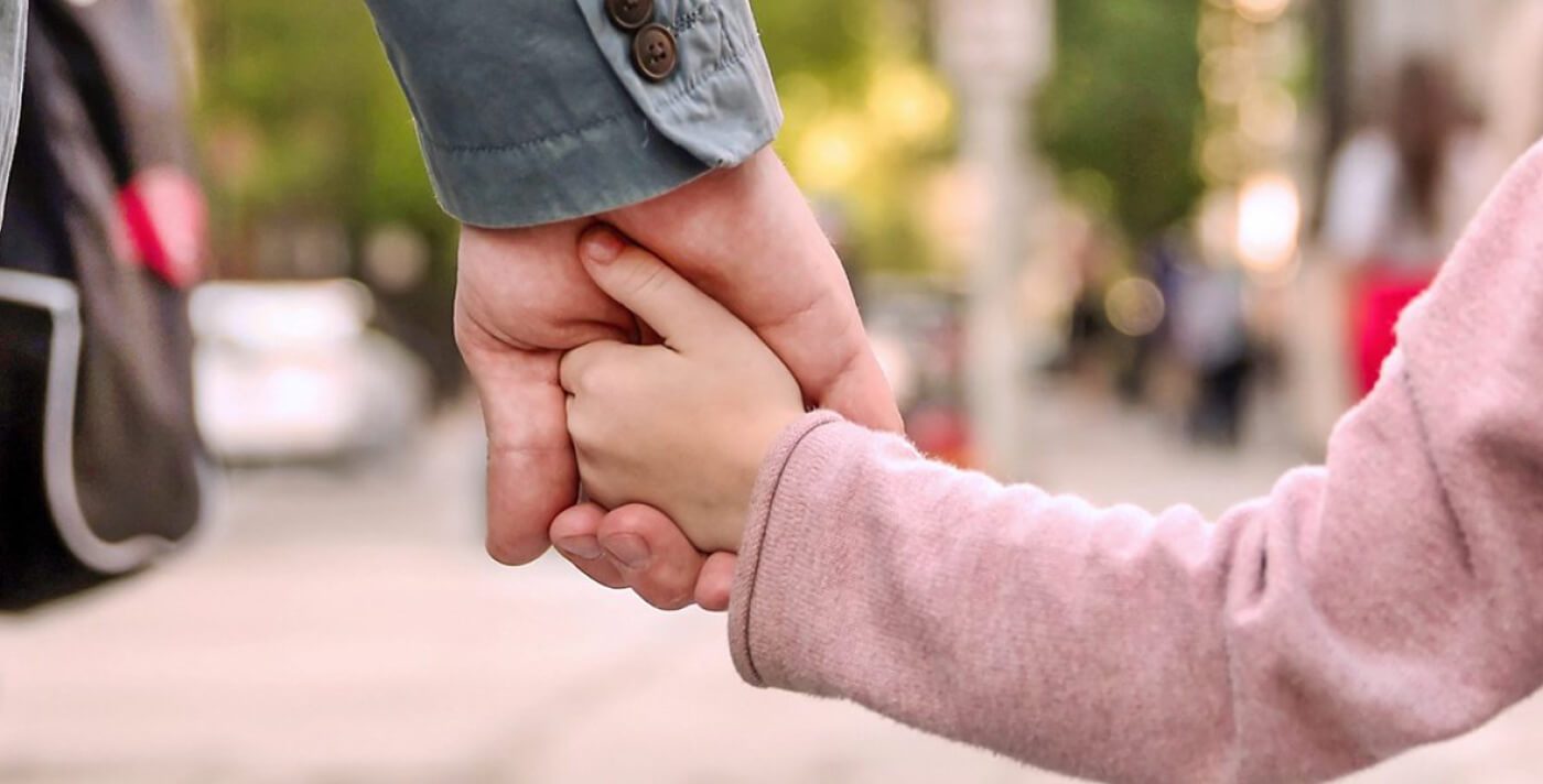 Stock/Familie Das Bild zeigt eine Szene, in der ein Erwachsener eine junge Person oder ein Kind an der Hand hält. Die Hände der beiden Personen sind mit emotionaler Wärme und Sicherheit verbunden; dies symbolisiert Schutz, Fürsorge und Vertrauen. Der Hintergrund deutet auf eine städtische Umgebung hin, möglicherweise auf einem Bürgersteig oder einer belebten Straße. Die Kleidung der Personen, ein Jackett und bequeme Kleidung, verstärkt den Eindruck von Alltagssituationen und familiärer Nähe.